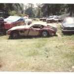 Classic car in a junkyard, possibly an MG, surrounded by overgrown vegetation and other scrap, hinting at neglect and disuse.