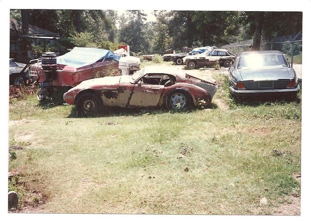 Classic car in a junkyard, possibly an MG, surrounded by overgrown vegetation and other scrap, hinting at neglect and disuse.